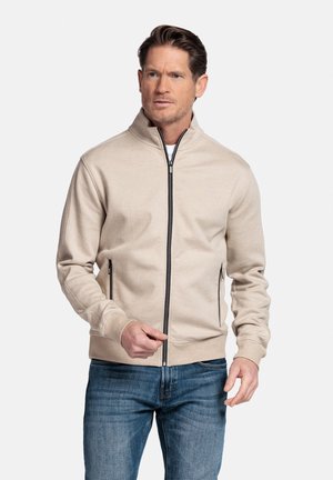 Man draagt een beige jas met ritssluiting in het zwart en een blauwe spijkerbroek, staand tegen een effen witte achtergrond.