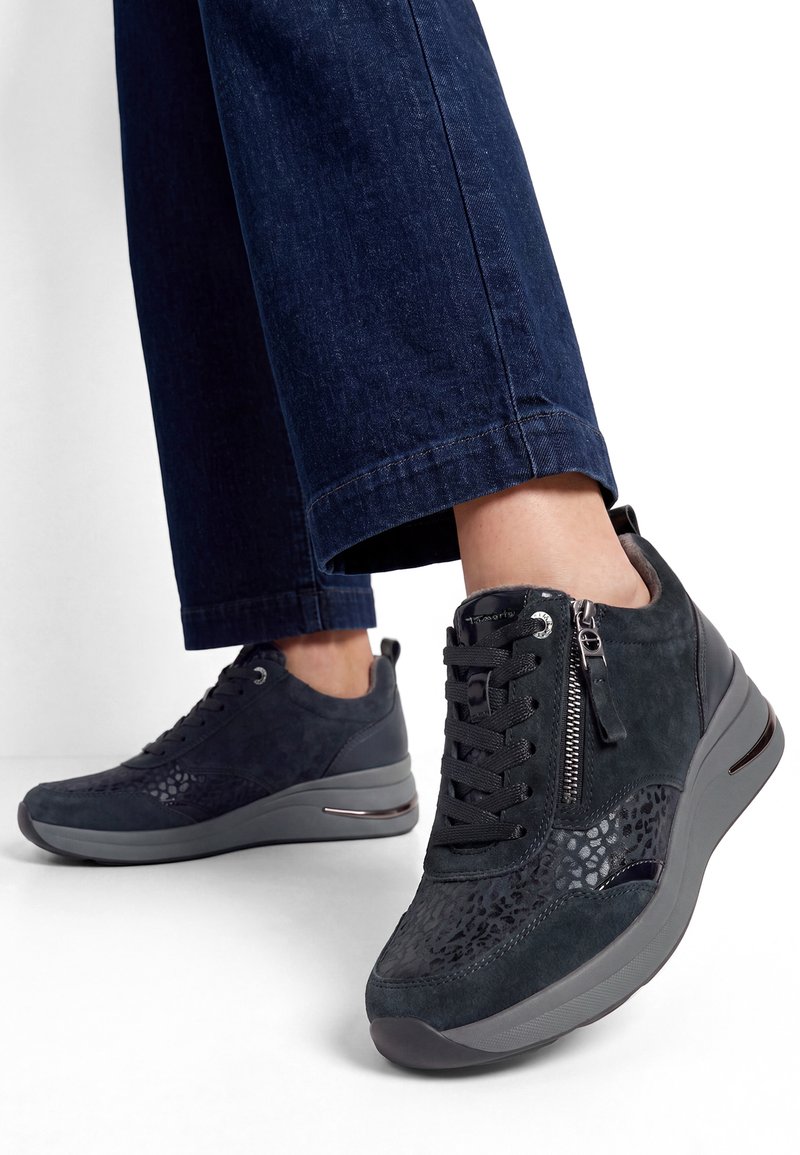 Persona con vaqueros azul oscuro y zapatillas negras de cuña con cordones y cremallera lateral, de pie sobre fondo blanco.