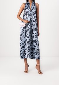 Femme portant une robe midi sans manches à motif bleu et noir, des sandales à talons hauts beiges et un petit sac bandoulière rose.