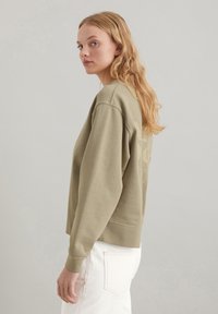 Olivgrüner Pullover mit lockerer Passform, gerippten Bündchen und abgerundetem Saum. Verfügt über dezente Logo-Details auf der Rückseite. Kombiniert mit weißen Jeans.