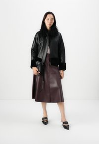 Veste en cuir noir avec col en fourrure, jupe midi en cuir marron, chaussures noires à bout pointu et petit sac à main noir avec une bandoulière.