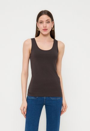 Młoda kobieta z prostymi brązowymi włosami, nosząca złote kolczyki-koła, dopasowany czarny top bez rękawów i niebieskie dżinsy, stojąca na białym tle.