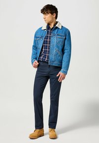 Chaqueta de mezclilla con cuello de borreguillo, color azul, dos bolsillos en el pecho; combinada con pantalones azul marino y botas de gamuza marrón claro con cordones.