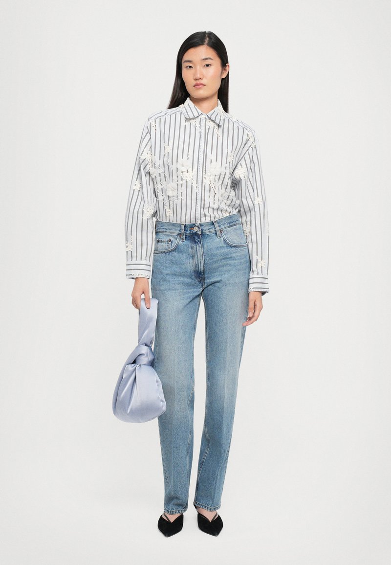 Prugasta bela in siva srajca s cvetličnimi vezeninami, v kombinaciji z modrimi ravnimi jeans hlačami in svetlo modro vozlano torbico. Črne konične čevlje.