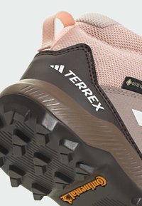 Smešasti smešasti planinarski cipele smeđe i ružičaste boje s gornjim dijelom od mrežaste tkanine, gumeni potplat s dubokim reljefom i "TERREX" logotipom sa strane. Vidljiva oznaka vodootpornosti GORE-TEX.