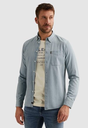Mann trägt ein hellblaues Hemd über einem cremefarbenen T-Shirt mit Grafik und blaue Jeans, steht vor einem einfarbigen Hintergrund und schaut nach rechts.