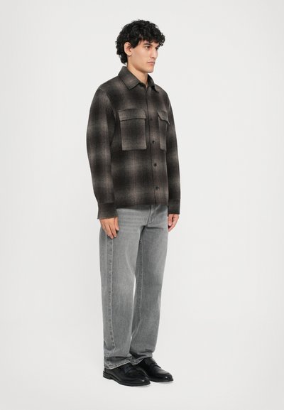 Man wearing a black and gray checkered button-up shirt with large chest pockets, gray jeans, and black dress shoes, standing sideways.