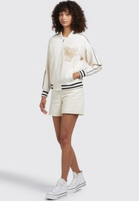 Vit bomberjacka med guldigt tigeremblem, svarta ränder på ärmsluten, i kombination med beige shorts och vita högtoppssneakers.