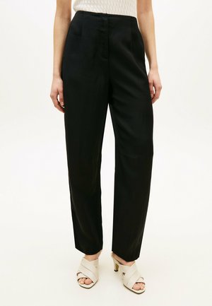 Donna che indossa pantaloni neri a gamba dritta a vita alta abbinati a sandali bianchi con tacco e un top bianco senza maniche dalla texture.