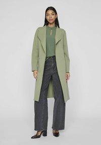 Light green wrap coat with a shawl collar, paired with a dark green top and high-waisted wide-leg jeans; model wears dark heels.