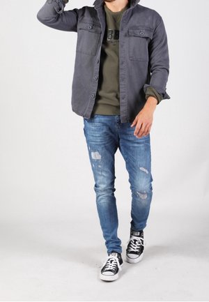 Homme marchant portant une veste grise boutonnée, un pull vert, un jean bleu déchiré et des baskets noir et blanc sur fond blanc.