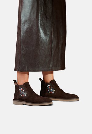 Bottes en daim brun foncé montant à la cheville avec broderie florale portées avec une jupe en cuir brun foncé.