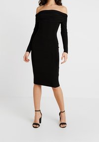 Žena ve černých šatech bez ramínek s dlouhým rukávem a délka ke kolenům, doplněná černými vysokými sandály na kotníku.