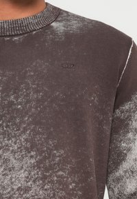 Pullover mit Rundhalsausschnitt in einem gesprenkelten Grau-Braun-Muster und einem kleinen, ovalen, auf der Brust gestickten Logo.