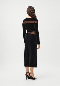 Melna samta kleita ar garām piedurknēm, caurspīdīgu augšdaļu un pieguļošu dizainu. Garums līdz pusei teļiem ar gludu tekstūru. Melni augstpapēžu apavi.