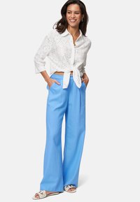 White eyelet blouse tied at the waist, paired with blue wide-leg pants. Model also wears white sandals, showcasing a relaxed, casual look.
