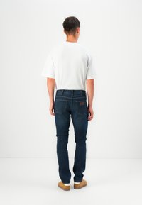 Jeans de denim azul escuro com um corte slim, apresentando bolsos traseiros e detalhes em couro. Usados com uma camisa de manga curta branca.