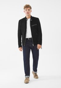Černá semišová bunda s kapsami na zip, bílý tričko pod tím, tmavé džíny a béžové boty. Rovný střih; casual design.