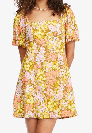 Kvinde iført en kort, gul blomsterkjole med pink og hvide blomster og puffede korte ærmer, stående med hænderne bag ryggen.