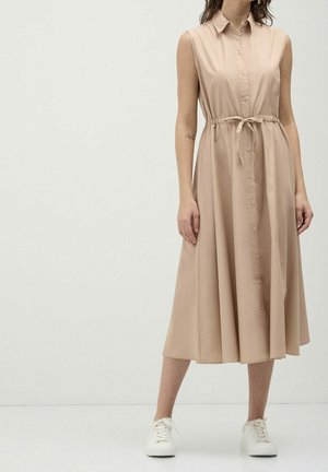 Kvinde iført en ærmeløs beige midi skjortekjole med knapper foran og bindebånd i taljen, kombineret med hvide sneakers, stående mod en hvid baggrund.