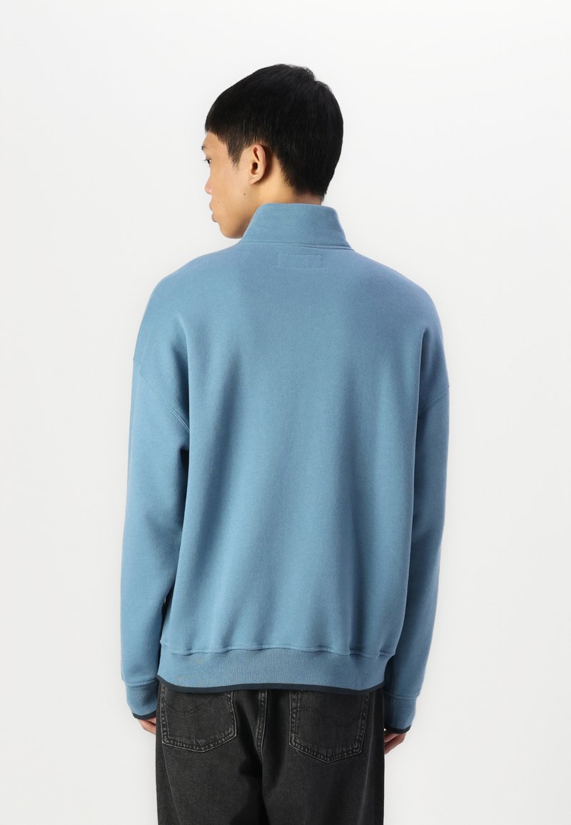 Lichtblauwe sweatshirt met een hoge kraag, geribde manchetten en een ontspannen pasvorm. Vlotte textuur, minimale merkaanduiding op de achterkant nabij de kraag.