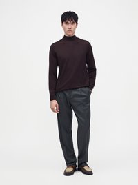 Homme debout portant un col roulé noir à manches longues, un pantalon gris et des chaussures marron et beige à enfiler sur un fond blanc uni.