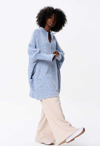 Lichtblauwe oversized gebreide cardigan met een rits aan de voorkant, geribde mouwen en zijsplitten, gecombineerd met crèmekleurige broek met streepjes en witte sneakers.