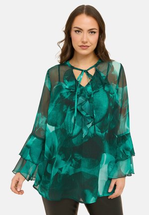 Kobieta ubrana w przezroczystą zieloną kwiecistą bluzkę z falbanianym wiązaniem przy dekolcie i warstwowymi dzwonowymi rękawami, stojąca na jednolitym tle.
