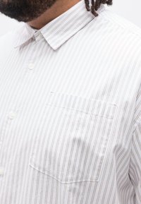 Άνδρας που φορά λευκό πουκάμισο με λεπτές κάθετες καφέ ρίγες και τσέπη στο στήθος, εμφανιζόμενος από το λαιμό έως το μέσο του κορμού.