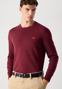 Mlad čovjek nosi usku bordo dugos kvornu majicu s malim crvenim "Hugo" logom, bež hlače i crni remen, s rukama u džepovima.