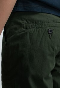 Pantaloni in tessuto leggero di colore verde con una texture liscia, dotati di chiusura con bottone e tasche laterali, visibili solo parzialmente nell'immagine.