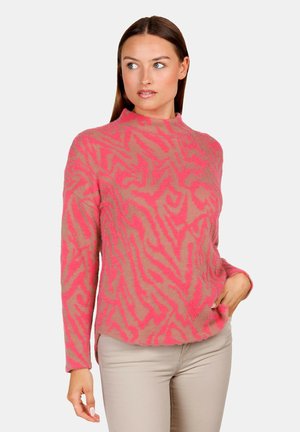 Frau trägt einen rosa-beigen Pullover mit Muster und langen Ärmeln sowie beige Hosen, schaut leicht seitlich vor einem einfarbigen Hintergrund.