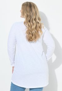 Žena s dlouhými vlnitými blond vlasy, oblečená v bílém tričku s dlouhým rukávem a modrých džínách, otočená zády k jednoduchému bílému pozadí.