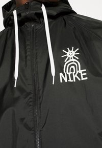 Černá lehká bunda s předním zipem a bílými šňůrkami, s grafickým designem slunce a duhy nad textem "NIKE."