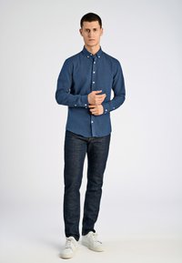 Man pastet de manchet van een donkerblauw overhemd met knopen, draagt donkere jeans en witte sneakers, staande tegen een effen lichte achtergrond.