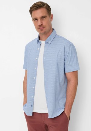 Man draagt lichtblauw kortärmelig overhemd over een wit T-shirt en rode broek, staat met handen in de zakken tegen een witte achtergrond.