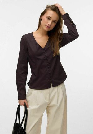 Femme portant un chemisier sombre à col en V et un pantalon plissé clair, tenant un sac à main noir, une main touchant ses cheveux.