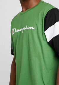 Camiseta de algodón verde con una manga negra y detalles blancos, que muestra el logo "Champion" en blanco en el pecho.