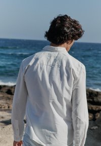 Chemise blanche à manches longues avec col boutonné, vue de dos. Le tissu semble léger. L'arrière-plan montre l'océan et une côte rocheuse.