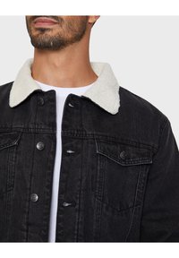 Zwarte denim jack met een witte, fluffy kraag, voor knoopsluiting en borstzakken; textuurstof met lichte gebruikssporen.