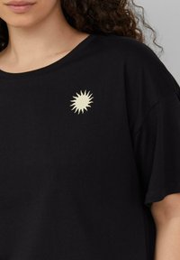 Schwarzes T-Shirt mit kurzen Ärmeln, das auf der linken Brust eine cremefarbene Sonnengrafik zeigt. Weicher Stoff mit lockerer Passform.