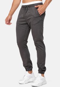 Grå joggingbukser med elastik i taljen, snøre og sidelommer; har en indsnævret pasform og ribbet kant. Bæres med hvide sneakers.