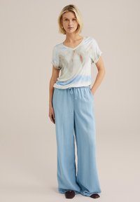 Lahki, majica iz viskose v tie-dye slogu pastelnih barv, v kombinaciji s širokimi hlačami svetlo modre barve, ki imajo elastičen pas.
