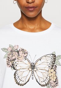 White t-shirt featuring a large butterfly graphic with detailed black outlines and pastel flower accents on one side. Short sleeves, round neckline.