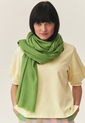 Žena s tmavými vlasmi a ofinou nosí svetlo žltý top a veľký jasne zelený šál prehodený okolo krku a ramien.