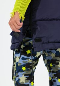 Mörkblå pufferväst med dragkedja och kardborrestängning, över mönstrade kamouflagedestinationer med blå och gröna toner samt gula stjärnor.