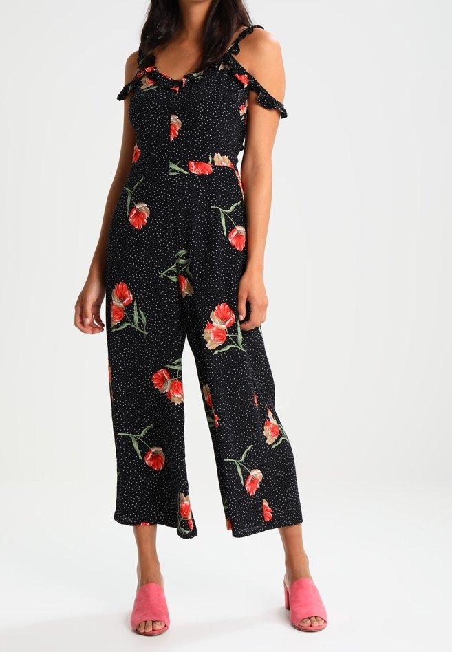 Czarny kombinezon w kwiatowy wzór z odkrytymi ramionami, w kropki oraz falbaniastymi akcentami. Modelka nosi różowe sandały na obcasie z odsłoniętymi palcami dla kontrastu.