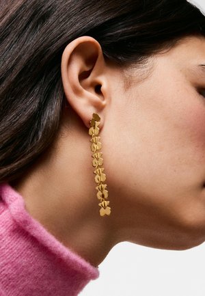 Primo piano dell’orecchio di una persona che indossa un lungo orecchino pendente d’oro fatto di forme di fiori collegati, con capelli scuri e un colletto di maglione rosa visibile.