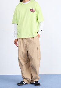 Grünes Baumwoll-T-Shirt mit kurzen Ärmeln und einem bedruckten Design auf der Brust, kombiniert mit beigen Cargo-Hosen und schwarzen Sneakers mit weißen Streifen.