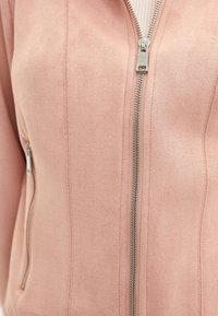 Różowa kurtka z ekoskóry zamszowej o teksturowanym wykończeniu, wyposażona w srebrny zamek błyskawiczny i boczne kieszenie. Pionowe przeszycia dodają detali.